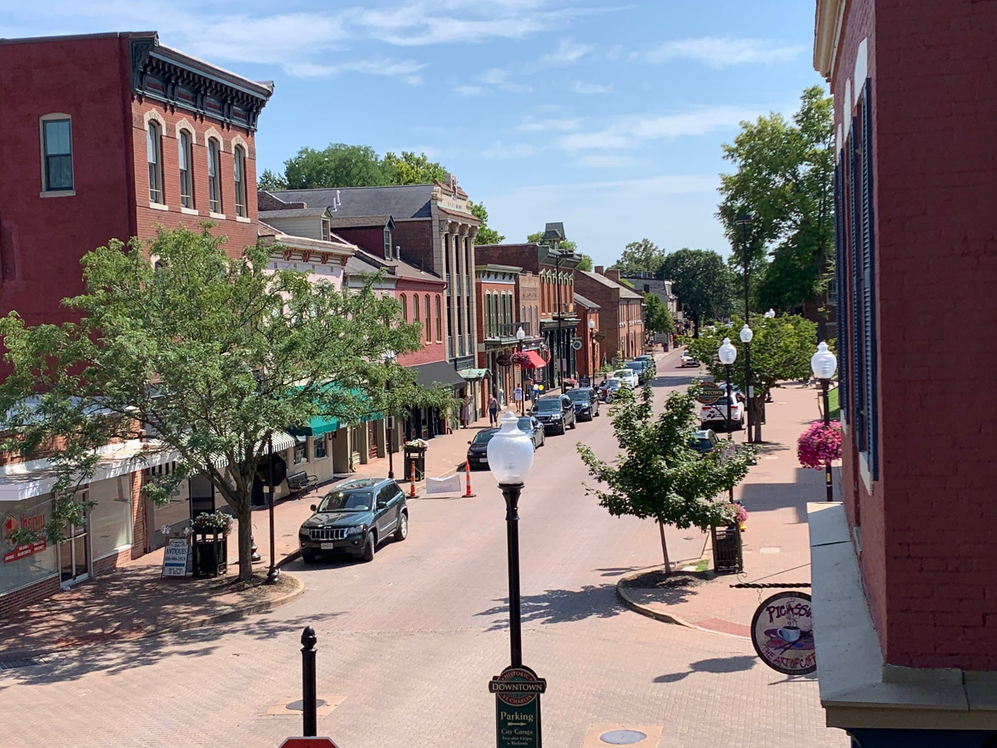 Small town street with brick buildings, parked cars, and trees on a sunny day.